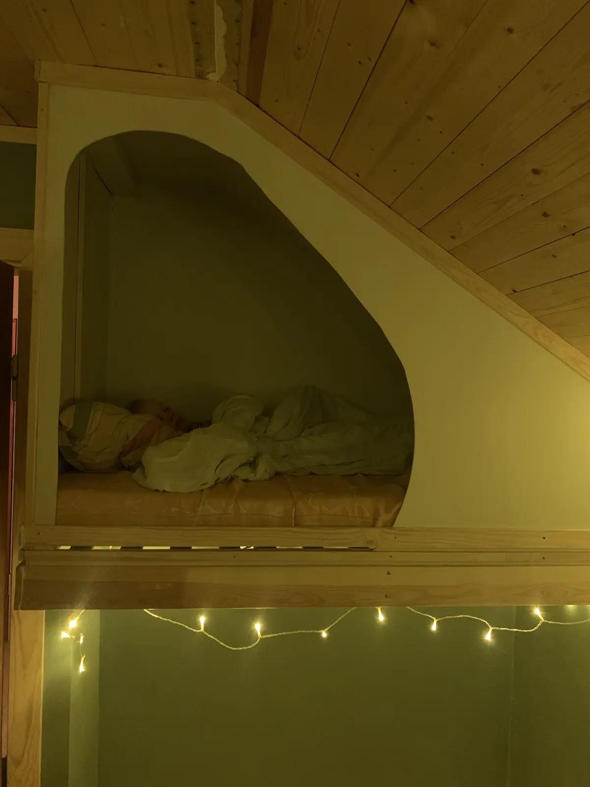 A person is sleeping in a small, arched alcove bed built into a wooden wall under a slanted ceiling. The bed has a mattress…