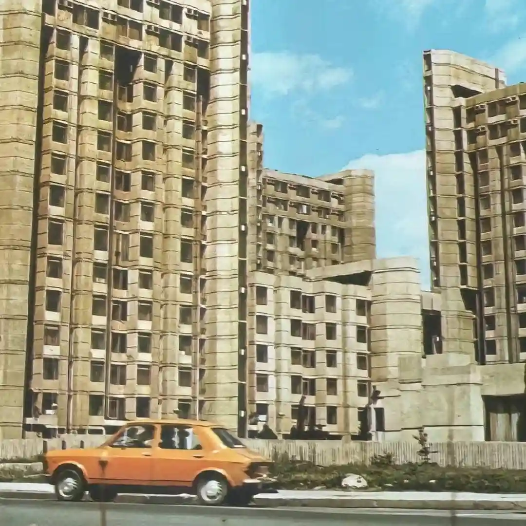 a vintage orange car drives past massive, unfinished concrete apartment blocks with many missing windows and exposed…