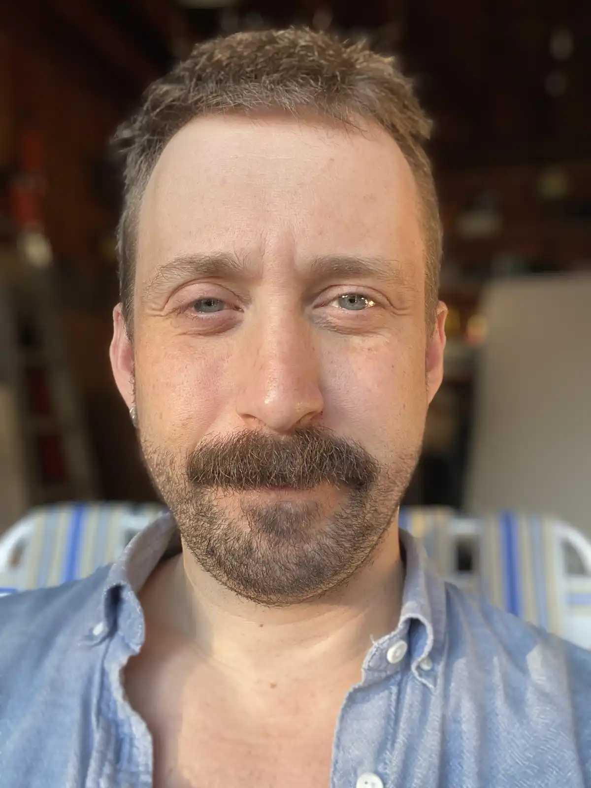 A close-up selfie of a man with a thick, dark mustache and stubble beard, wearing a light blue button-up shirt. He has short…