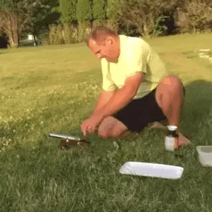 A man in a yellow shirt and black shorts sits on grass, preparing a small device. He points at it, then activates it…