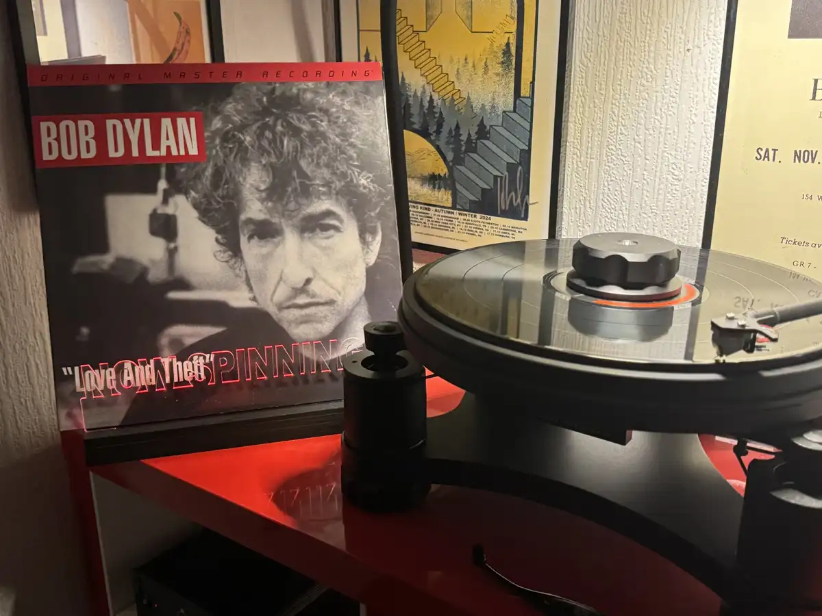 A vinyl record player sits on a red shelf with a Bob Dylan album titled 'Love And Theft' standing upright beside it. The…