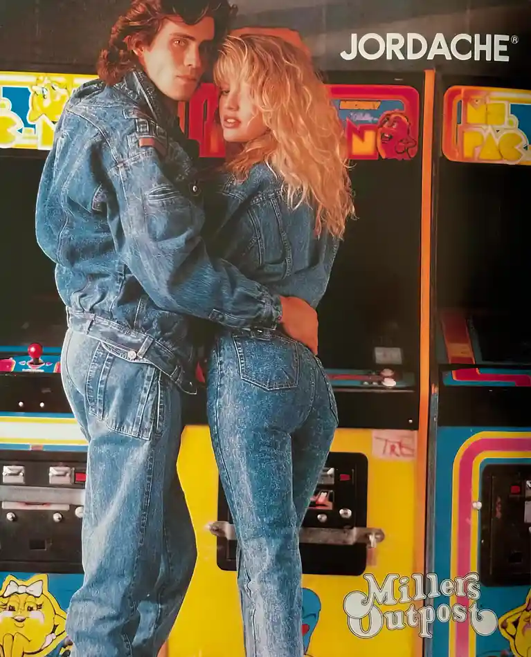 A vintage 1980s advertisement photo shows a man and woman in matching, baggy Jordache denim outfits hugging in front of…