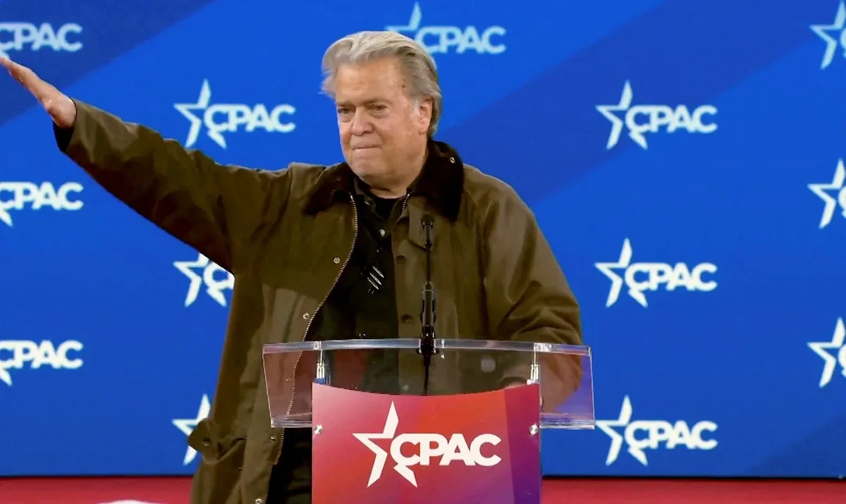 a man with gray hair wearing a brown jacket stands at a clear podium with a red CPAC logo, gesturing with his right arm…