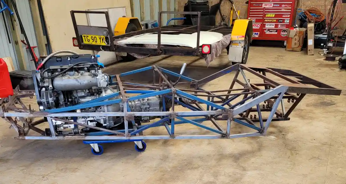 A partially assembled custom vehicle frame with an engine mounted on a wheeled cart sits in a garage, with a yellow trailer…