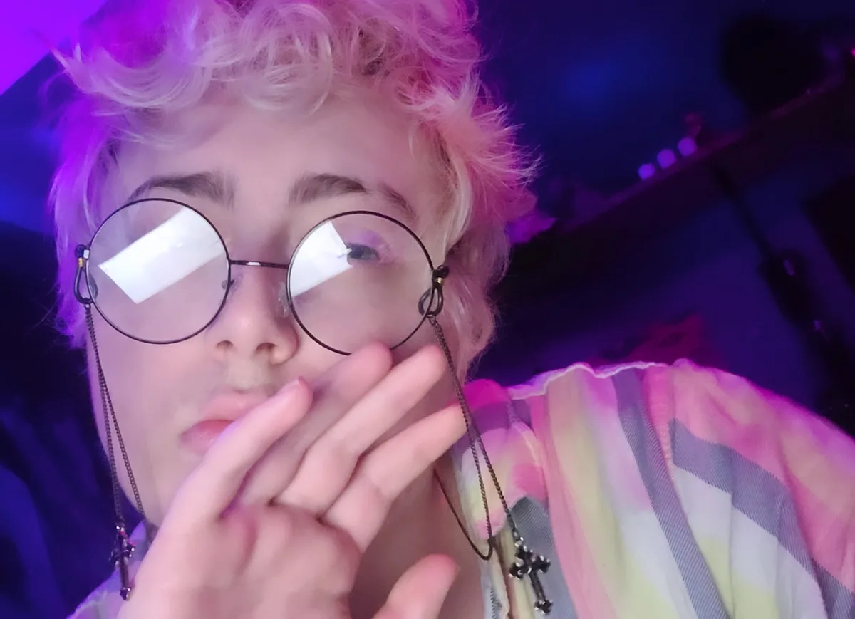 A person with curly blonde hair and round glasses, wearing a colorful striped shirt, covers their mouth with one hand while…