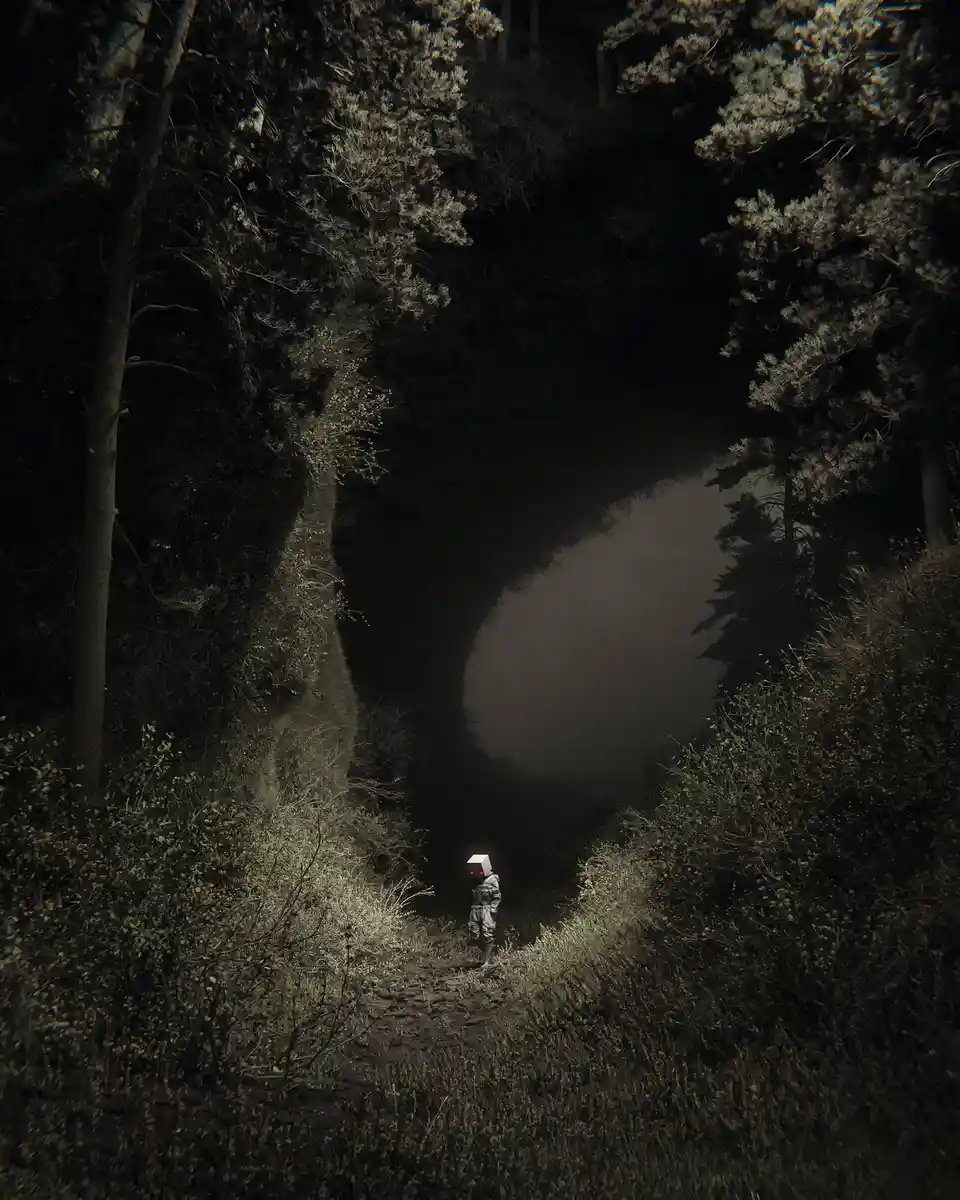A person wearing a white box-like helmet with glowing red eyes stands alone on a narrow dirt path in a dark, misty forest…