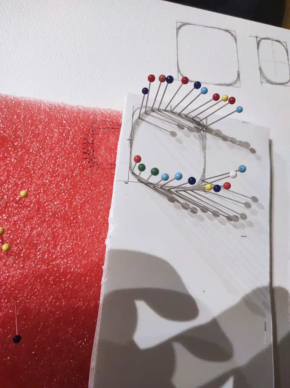 A sketch of a crown drawn on paper is pinned with colorful pushpins, with the shadow of a person holding a phone visible…