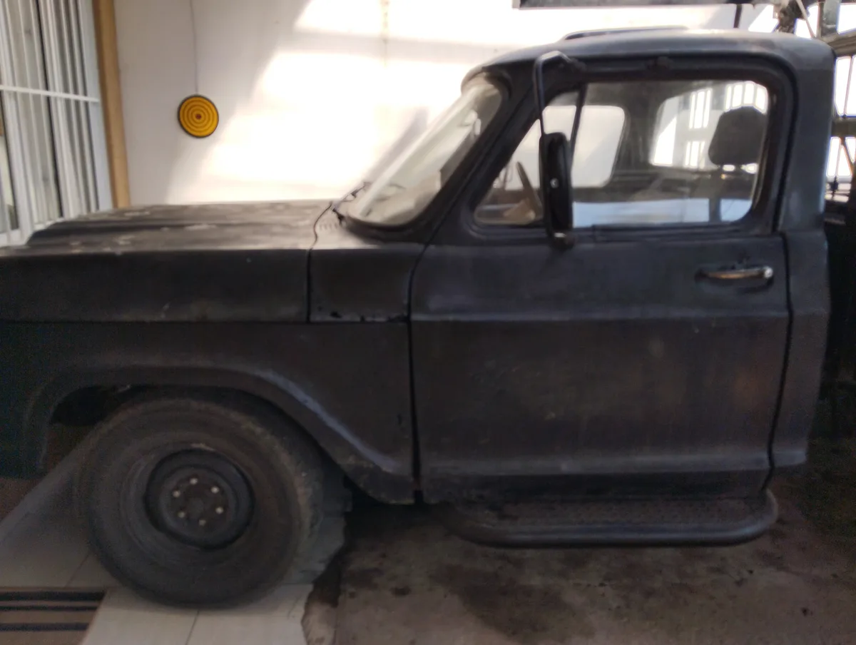 A black, rusty, old pickup truck is parked indoors, likely in a garage or workshop, with visible wear and dirt. The vehicle…