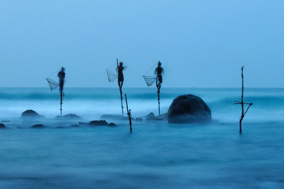 A long-exposure photograph shows three silhouetted stilt fishermen standing on poles in shallow ocean water, each holding a…