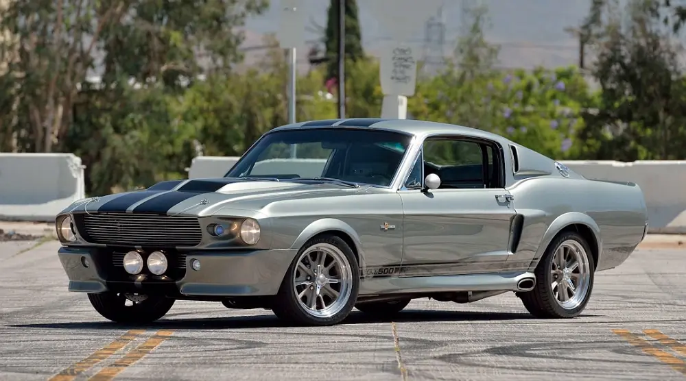 A silver classic Ford Mustang with black racing stripes and '427-1500' badging is parked on asphalt. The overlaid text reads…
