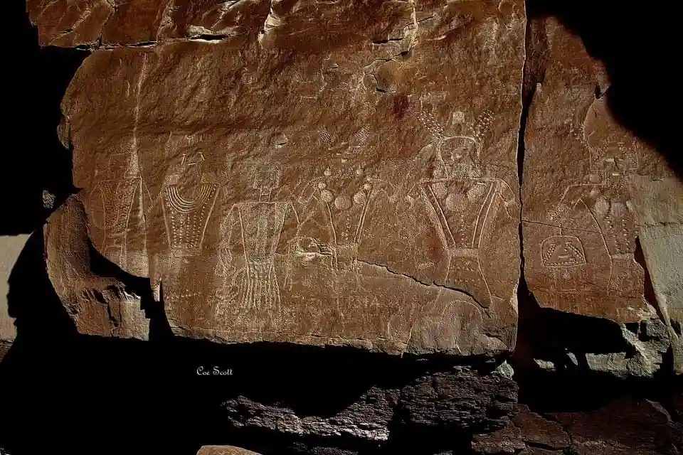 A photograph of ancient rock art etched into a large, weathered brown rock face, showing stylized human figures with…