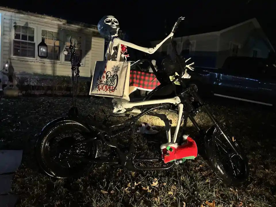 A skeleton decoration is posed on a black motorcycle in a yard at night, holding a tote bag that reads 'Joy to the World'…