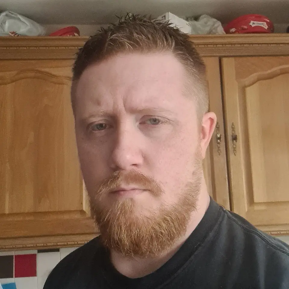 A man with red hair and a matching beard stares directly at the camera with a weary, unimpressed expression. He wears a…