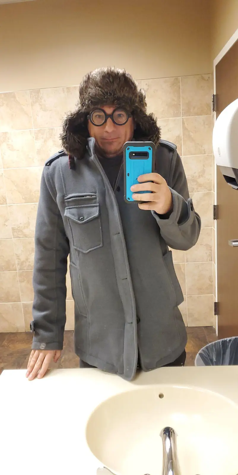 A man in a bathroom mirror selfie wearing a thick grey coat, a furry winter hat with ear flaps, and round glasses, holding a…
