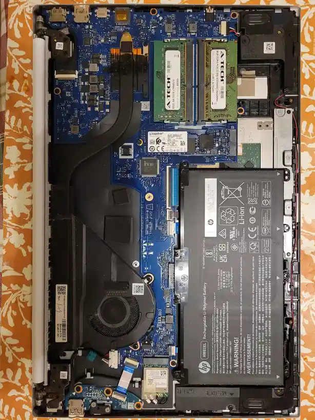 An overhead view of a disassembled laptop showing its internal components, including a blue motherboard, two RAM sticks, a…