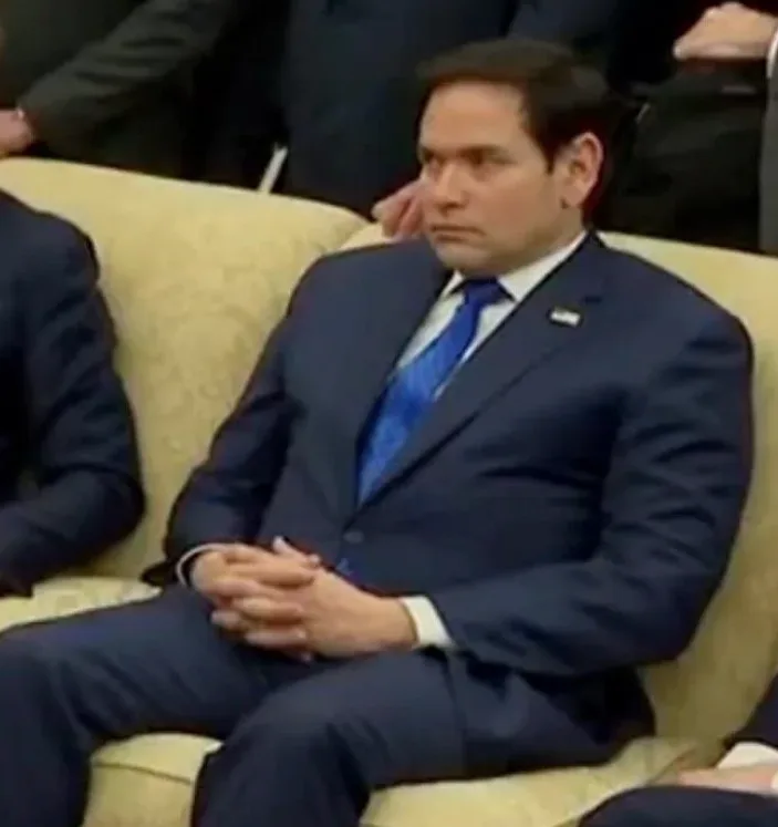 a man in a dark blue suit and blue tie sits on a beige couch with a bored, unimpressed expression, hands clasped in his lap…