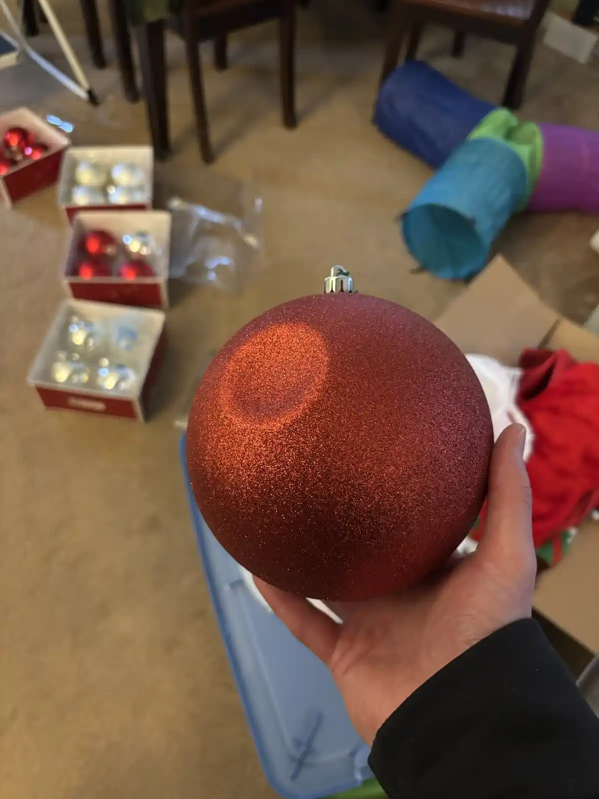 A hand holds a large, glittery red Christmas ornament with a noticeable circular smudge or stain on its surface. In the…