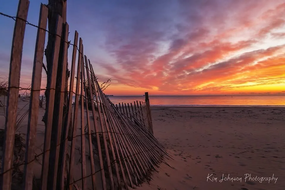 A serene beach sunset photo with a wooden fence in the foreground, featuring vibrant orange, pink, and purple clouds over…