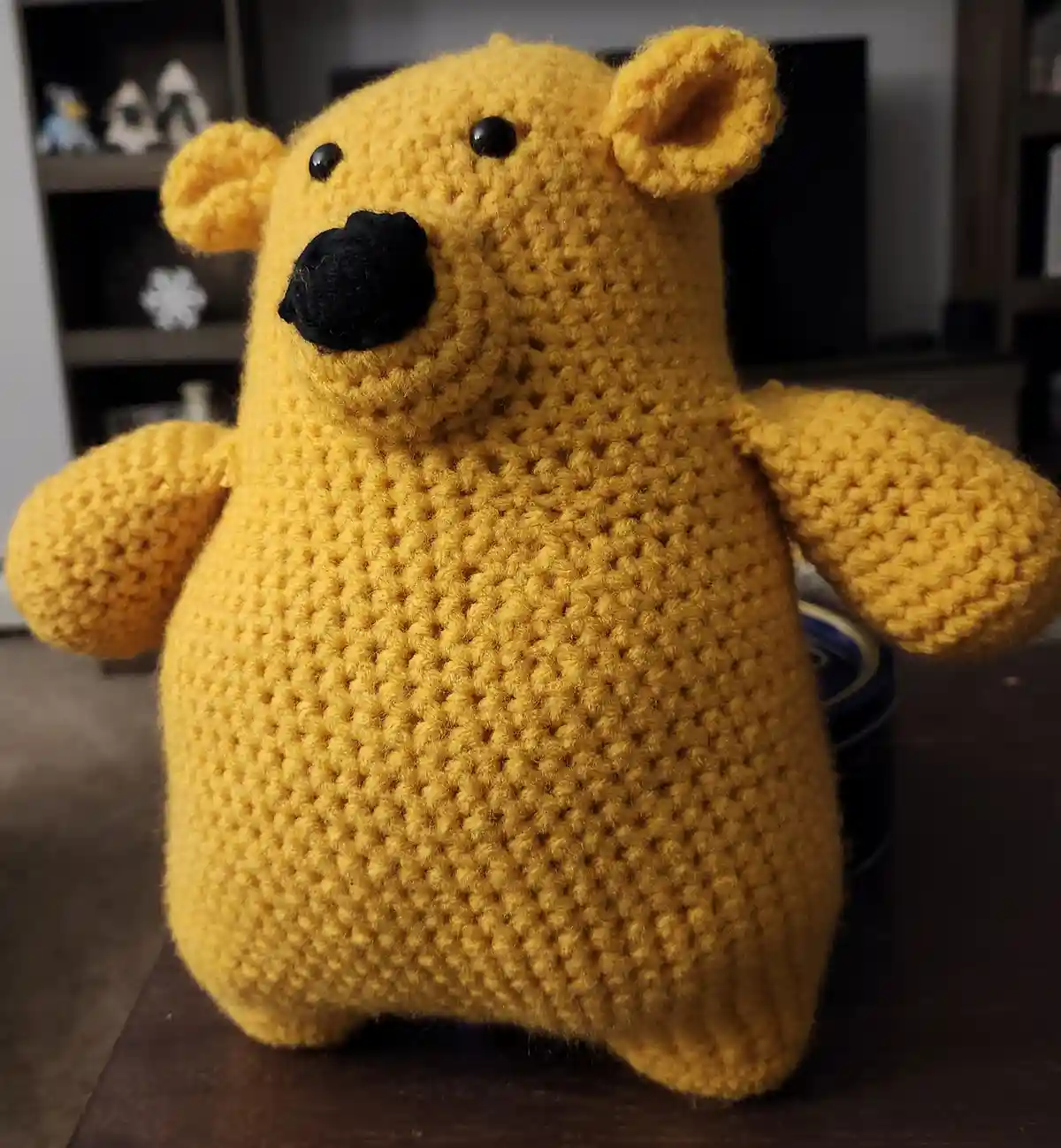A yellow crocheted bear with black button eyes and nose sits on a dark wooden surface. The background shows a shelf with…