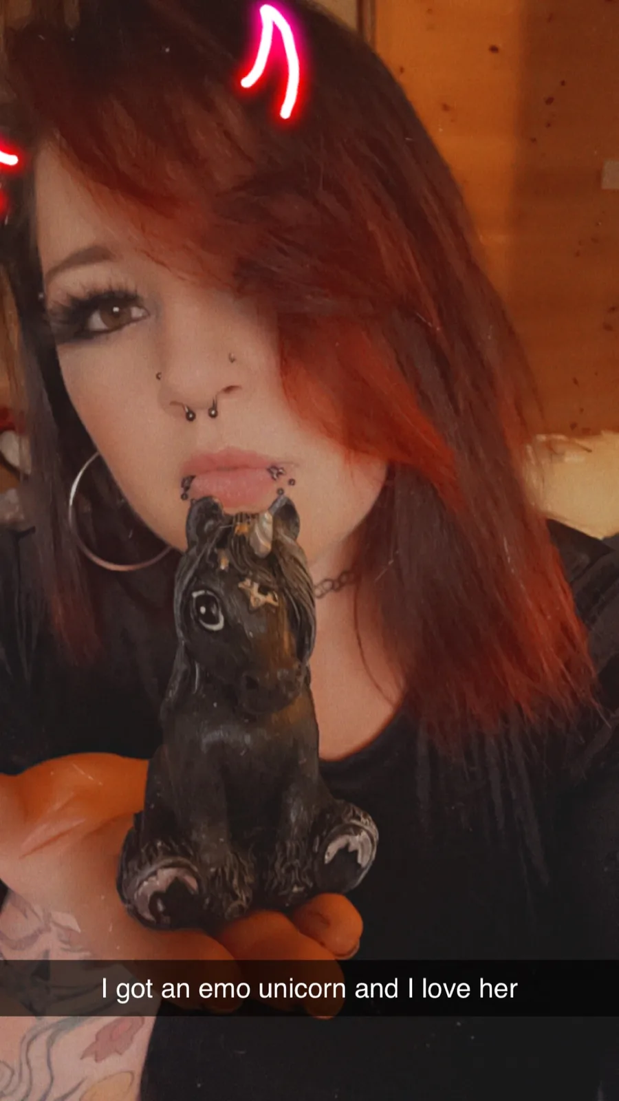 A person with red hair, multiple facial piercings, and neon pink devil horns holds a small black unicorn figurine. The…