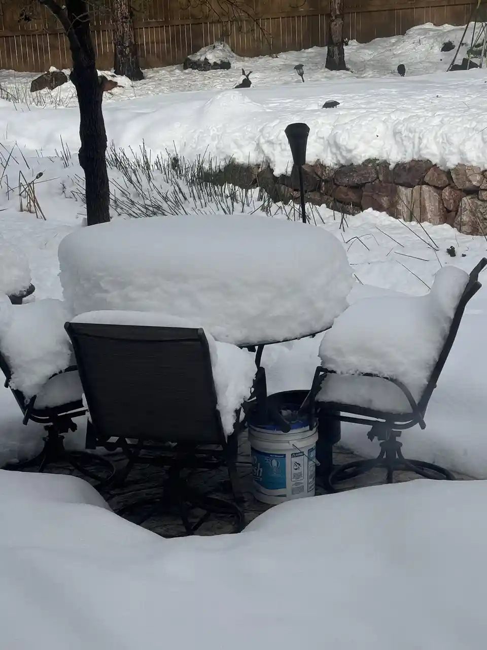 A backyard patio set, including a round table and several chairs, is completely buried under a thick layer of fresh snow. A…