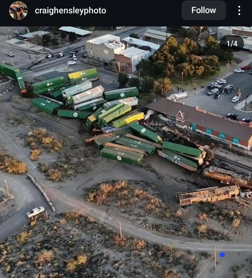 An aerial photo shows a massive train derailment with dozens of green and yellow freight cars scattered across a dirt lot…