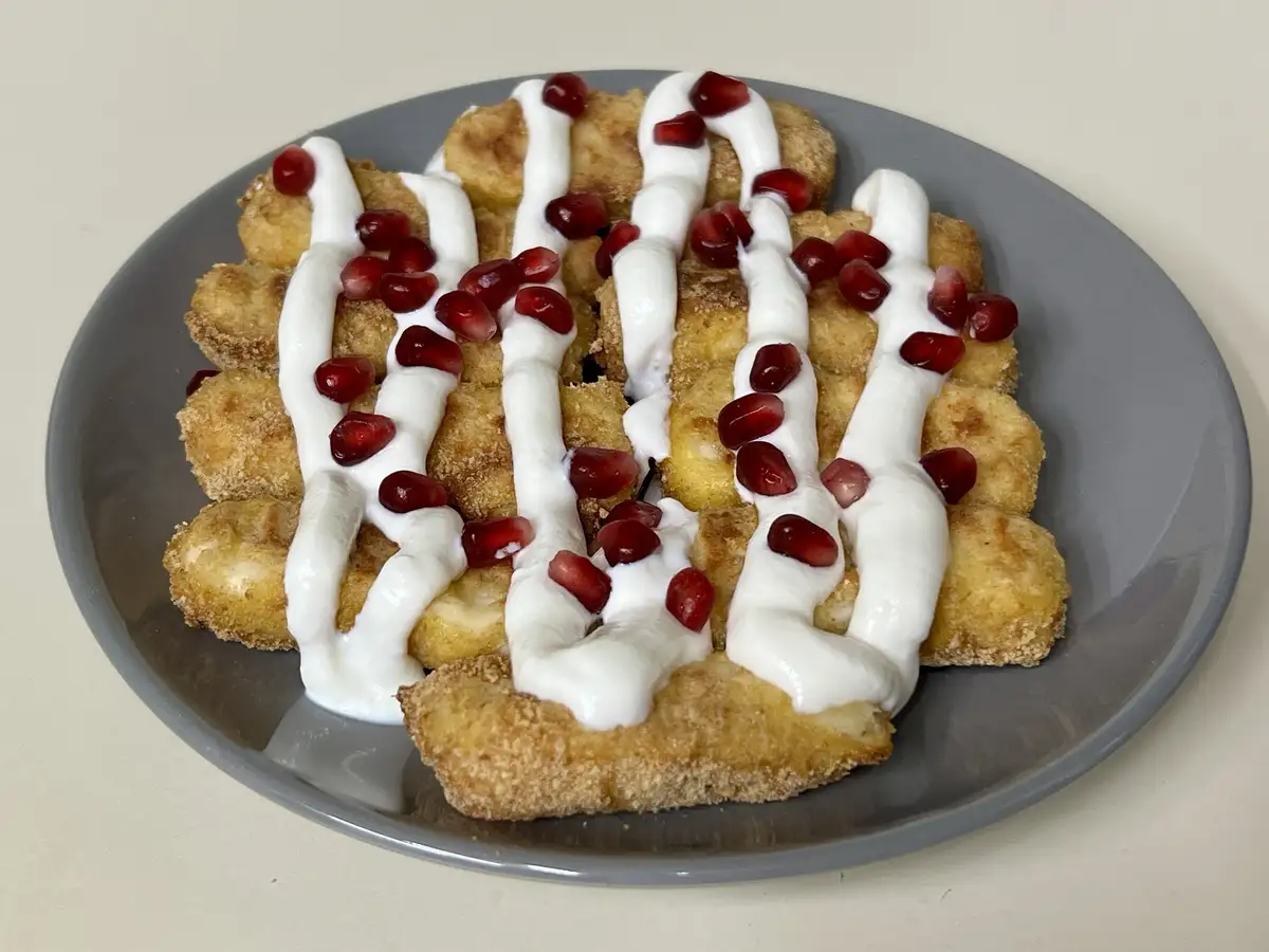 a gray plate holds golden-brown fried food pieces arranged in rows, drizzled with white sauce and topped with red…