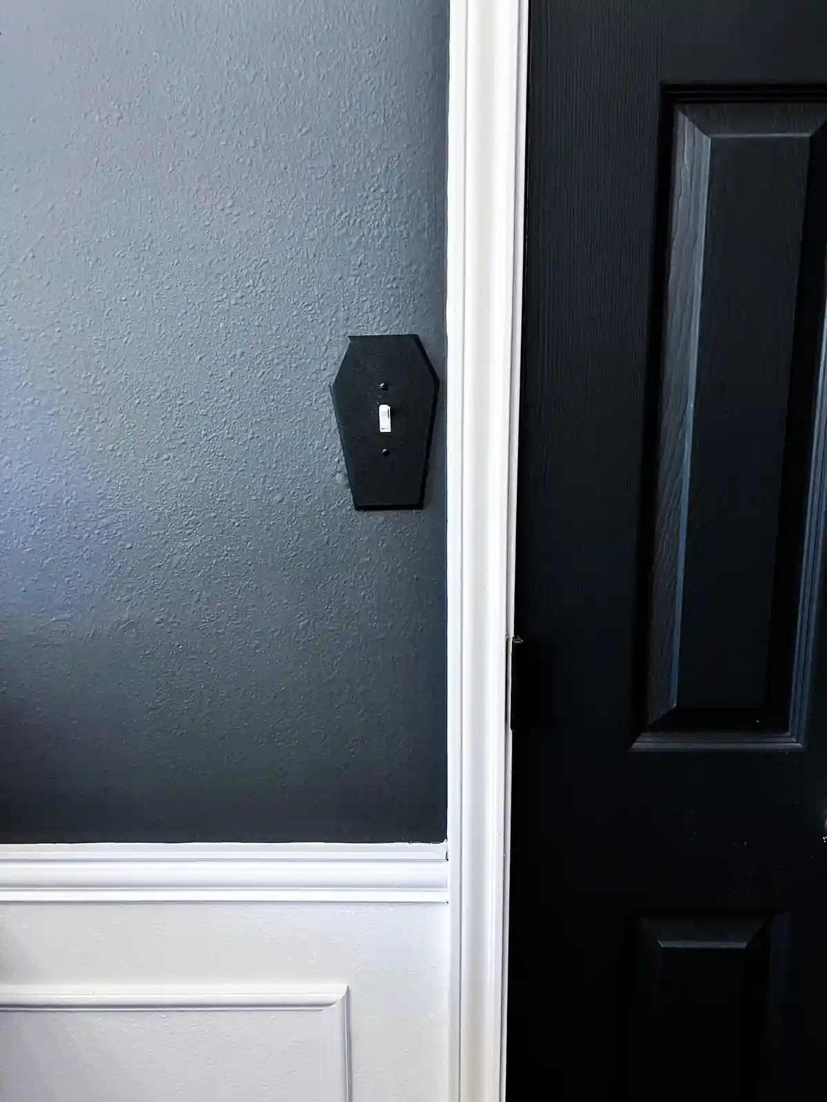A close-up photo shows a black light switch plate shaped like a coffin mounted on a dark gray wall next to a black door with…