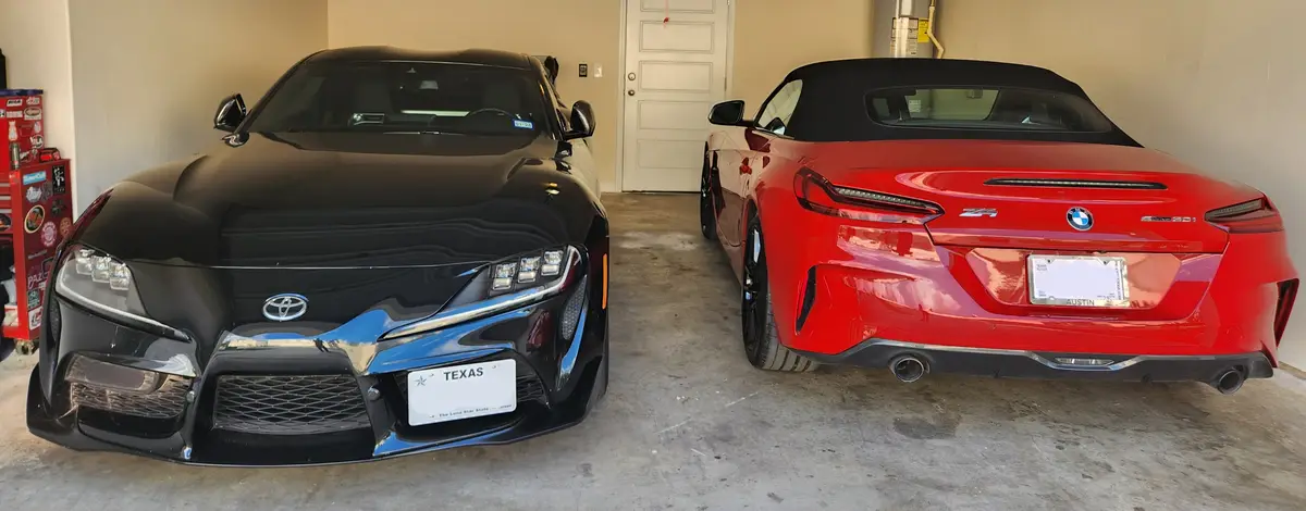 Two high-end cars are parked side-by-side in a garage: a black Toyota Supra facing forward on the left, and a red BMW Z4…