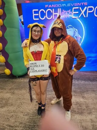 Two people in cosplay costumes pose at an event with a large sign that reads 'SHOW RESPECT TO ALL COSPLAYERS!!!'. One person…