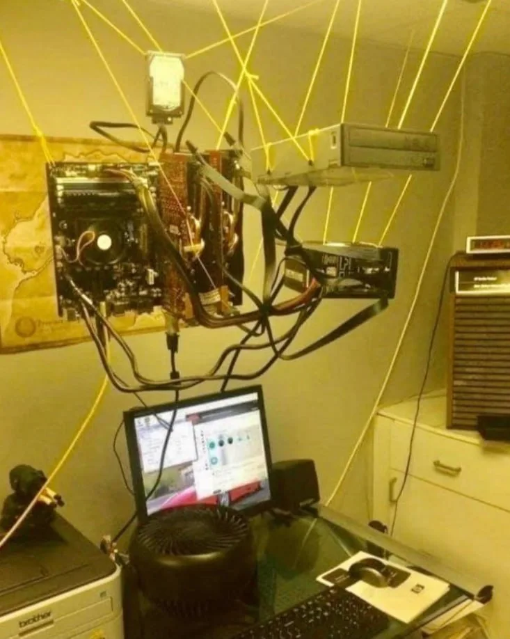 A chaotic, homemade computer setup hangs from the ceiling, connected by numerous yellow laser beams that shoot from its…
