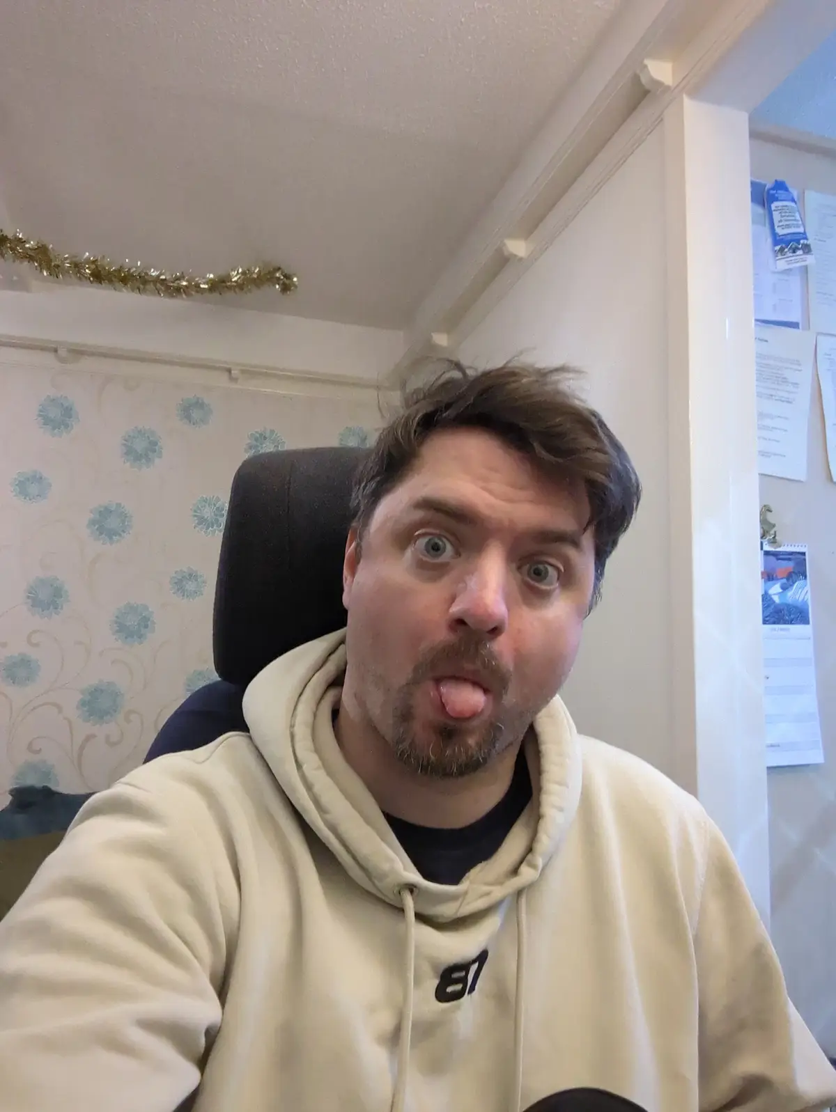 A man with brown hair and a goatee sits in a black chair wearing a beige hoodie, making a silly face by sticking his tongue…
