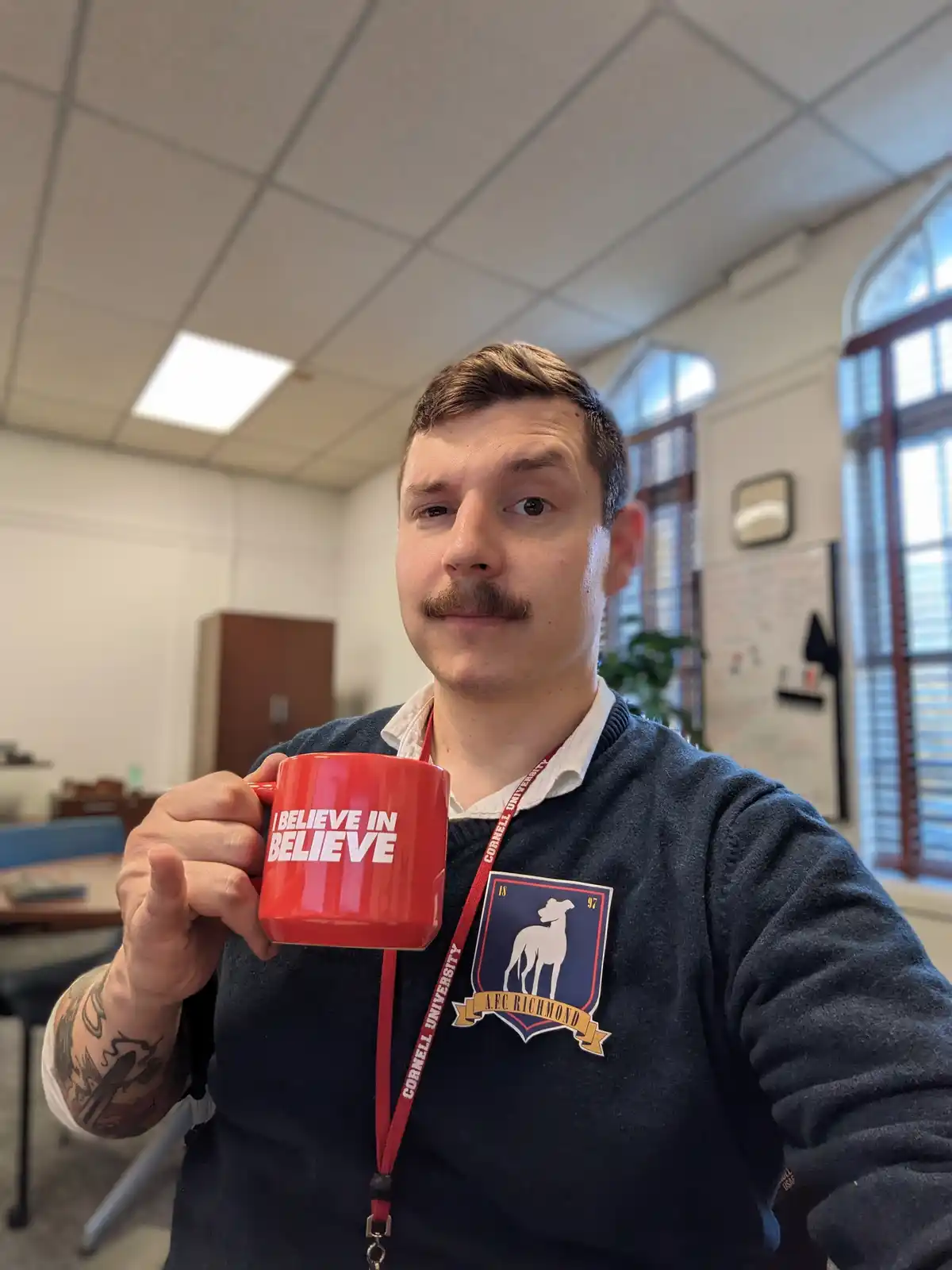 A man with a mustache holds up a red mug that reads 'I BELIEVE IN BELIEVE' in white text, creating a self-referential pun…