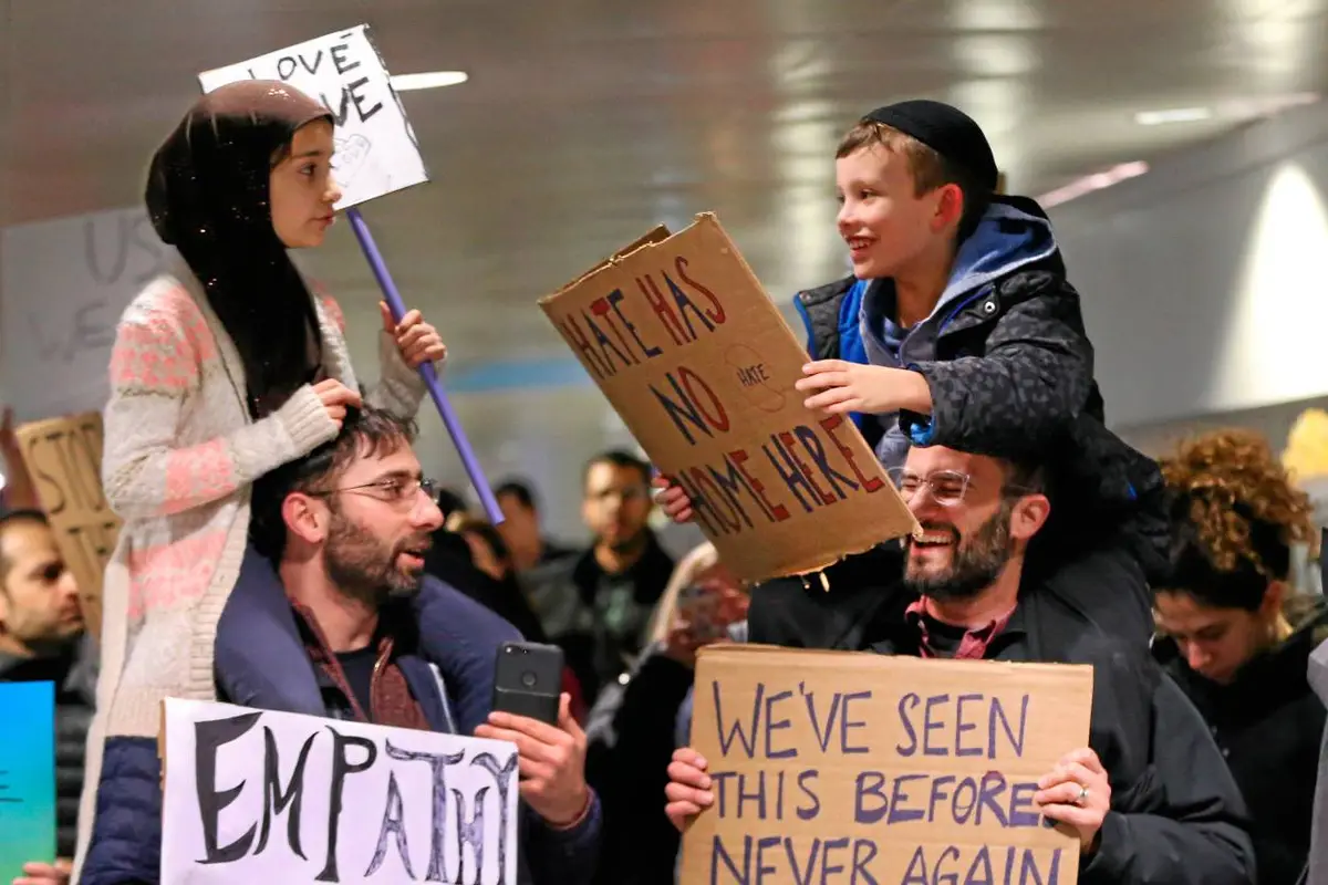 A photo shows two children at a protest, one holding a sign that reads 'HATE HAS NO HOME HERE' while sitting on an adult's…