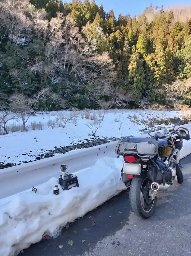 A motorcycle is parked on a snowy roadside next to a guardrail, with a small camping stove and mug set up on the snow. The…