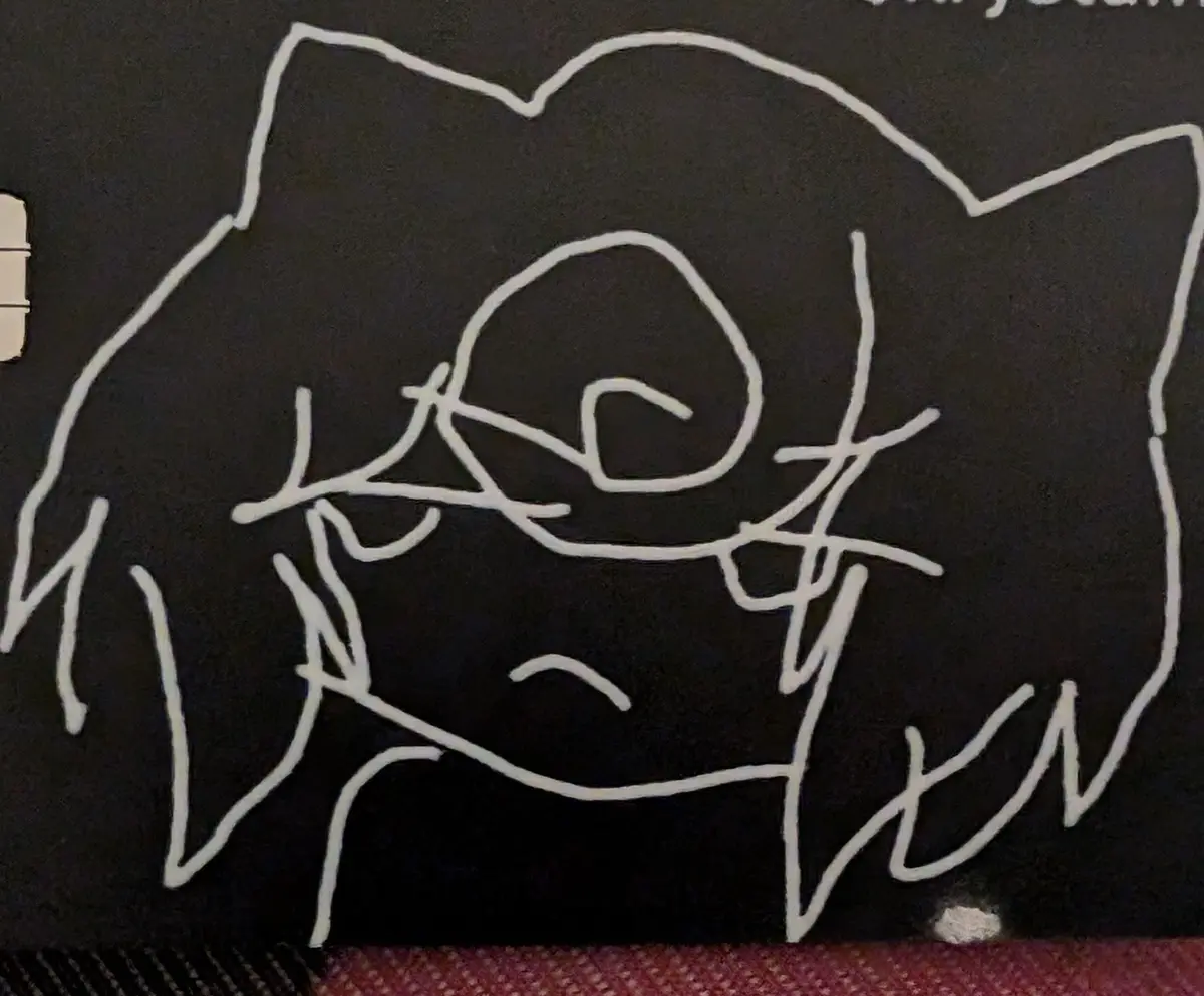 A crude white line drawing on a black surface depicts a stylized animal face with pointed ears, a snout, and a simple mouth…