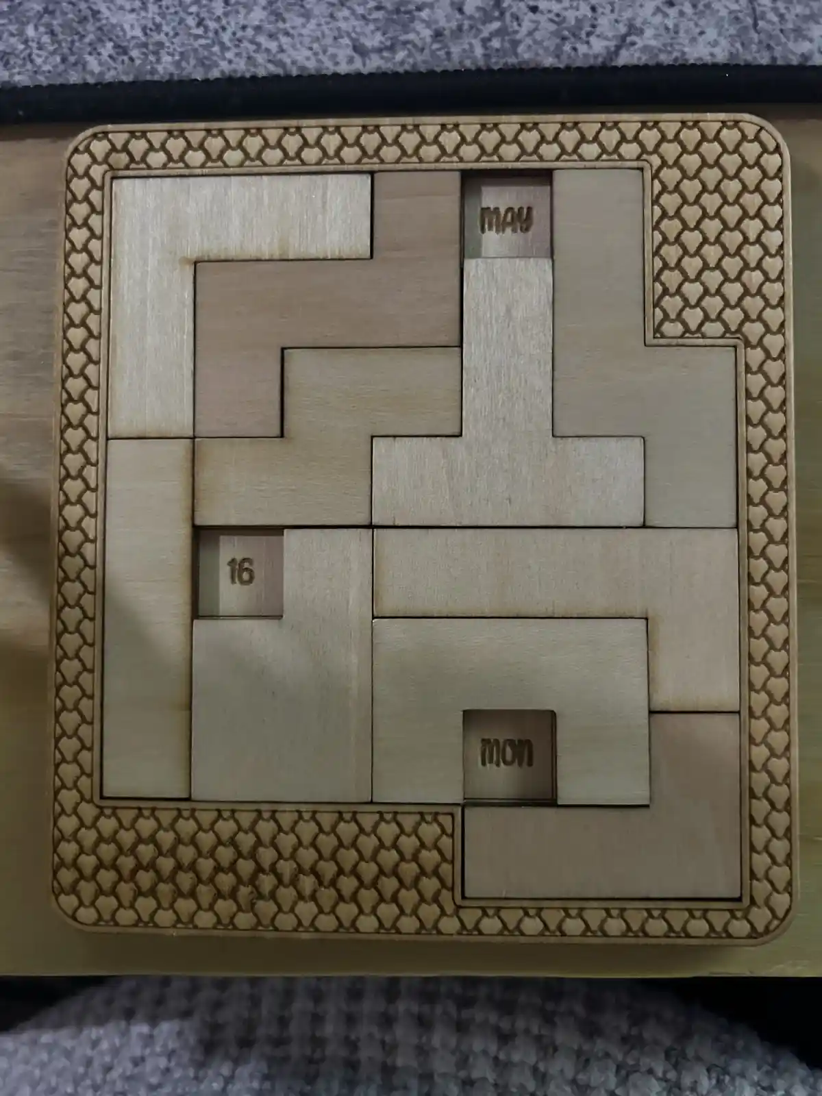A wooden puzzle calendar with interlocking pieces, showing the date '16' in a square, the month 'MAY' in another square, and…