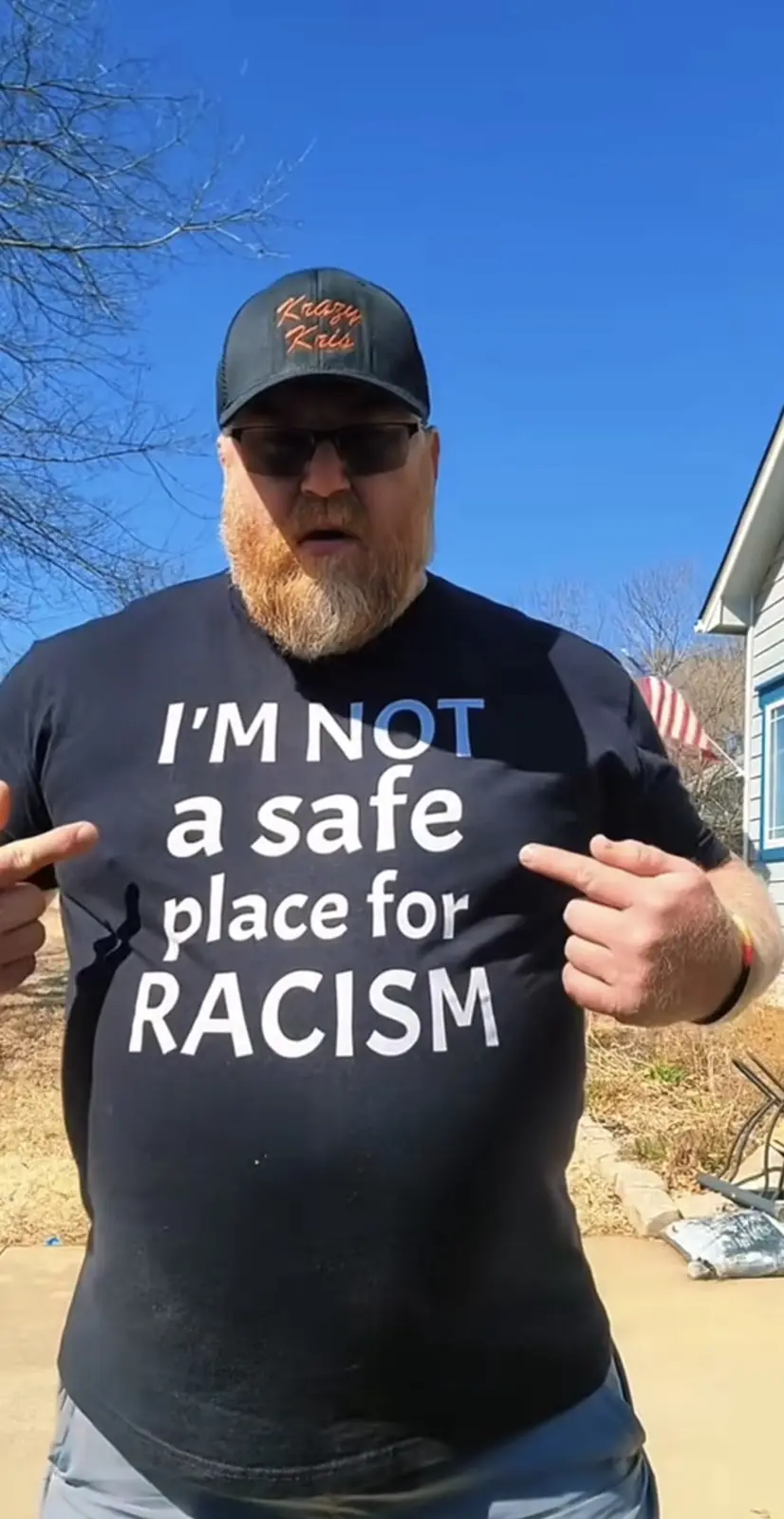 A man with a beard, sunglasses, and a black baseball cap stands outdoors in front of a house under a clear blue sky. He is…