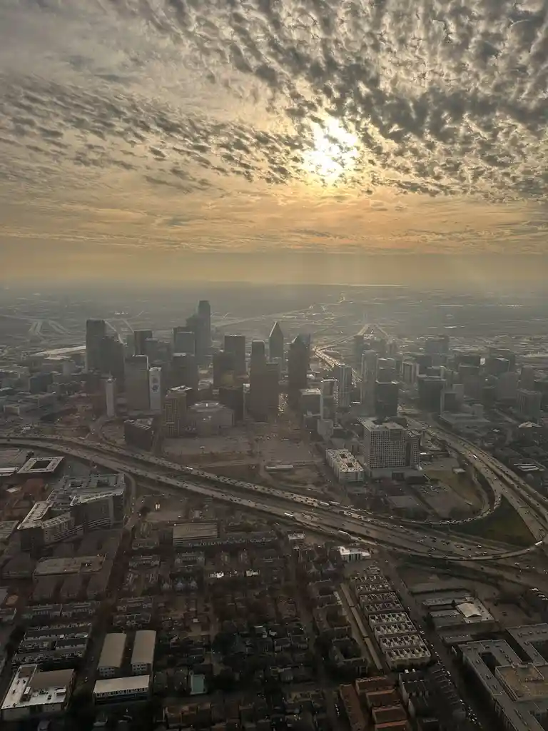 An aerial photo of a sprawling cityscape at sunrise or sunset, with golden light filtering through textured clouds above a…