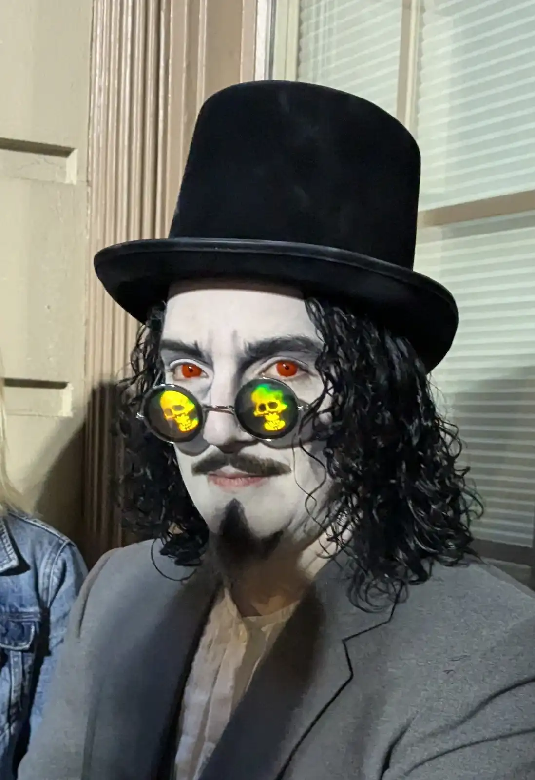 A person in a vampire costume with white face paint, orange contact lenses, a black top hat, and curly black hair, wearing…