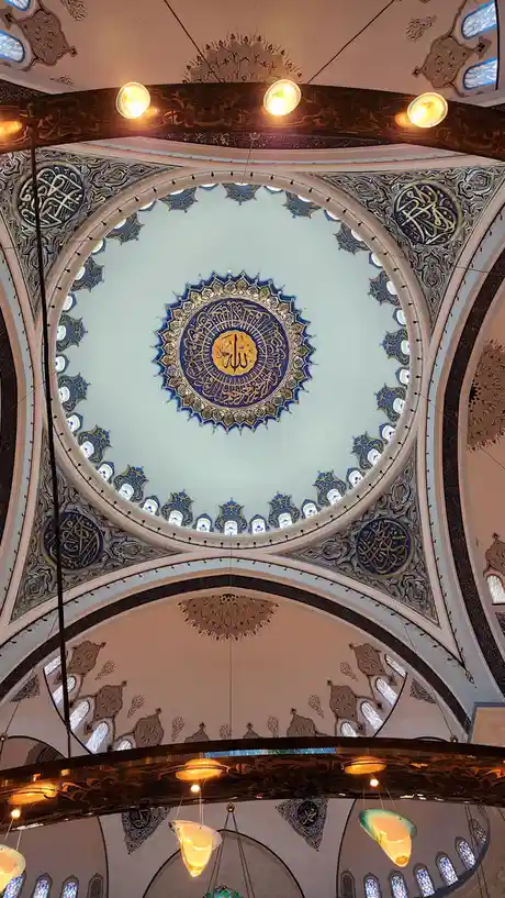 The image shows the ornate, domed ceiling of a mosque with intricate Islamic geometric and calligraphic patterns in blue and…