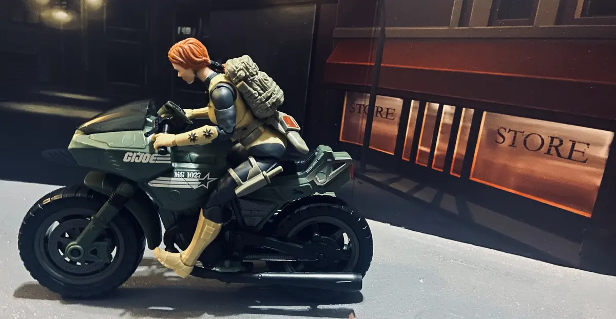 A G.I. Joe action figure with red hair and a tactical backpack is seated on a green motorcycle labeled 'G.I. JOE' and 'MG…