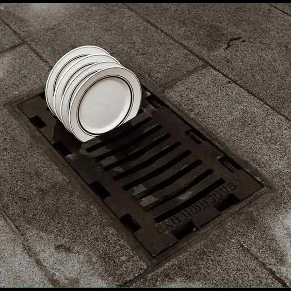 A stack of white ceramic plates with black rims sits precariously balanced on a metal storm drain grate embedded in a paved…