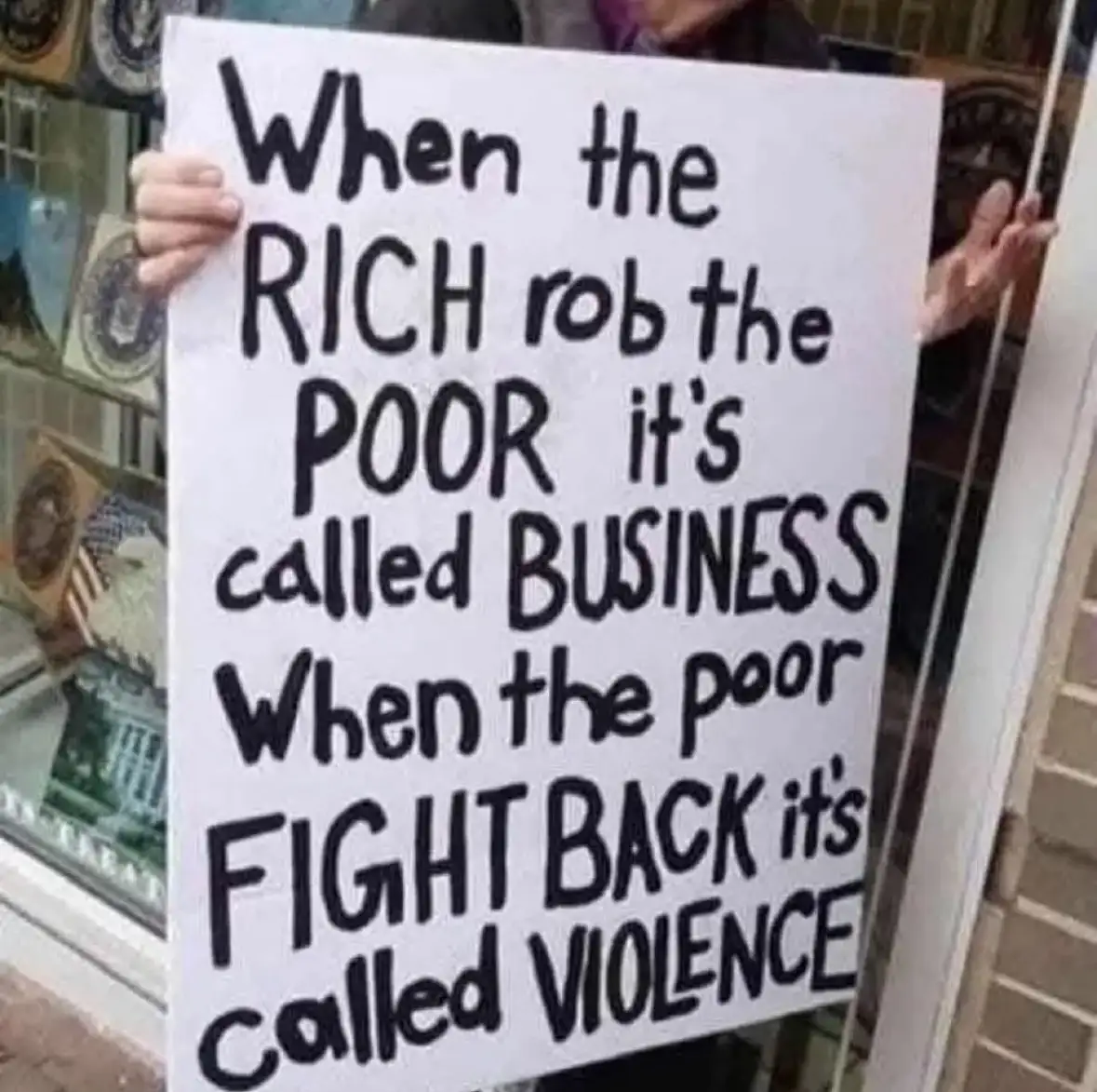 A person holds a white protest sign with bold, handwritten black text that reads: 'When the RICH rob the POOR it's called…