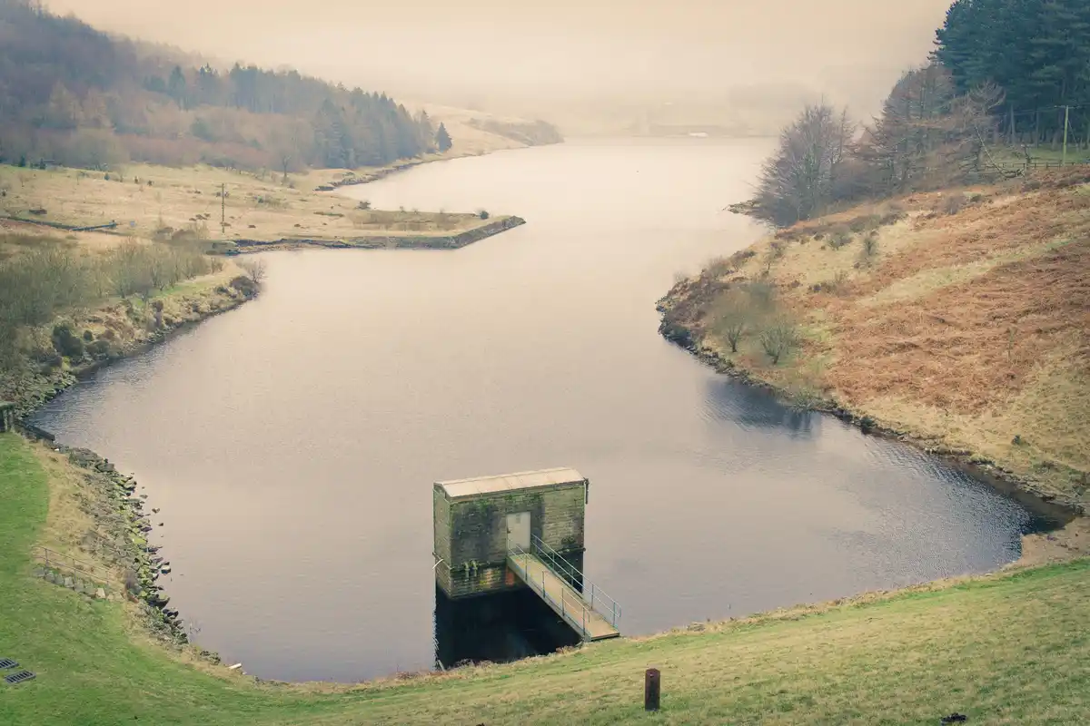 A misty, tranquil landscape featuring a calm reservoir surrounded by rolling hills and sparse trees. A small, isolated brick…
