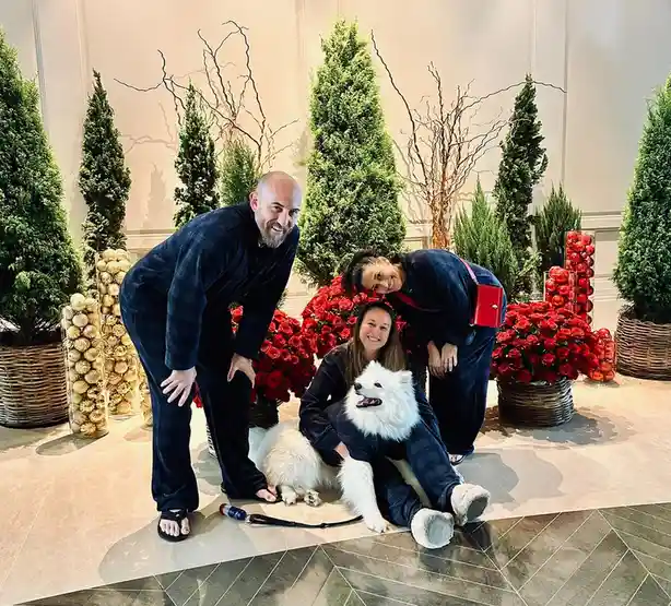 Three people and a fluffy white Samoyed dog pose together in matching dark plaid pajamas in a lavishly decorated indoor…