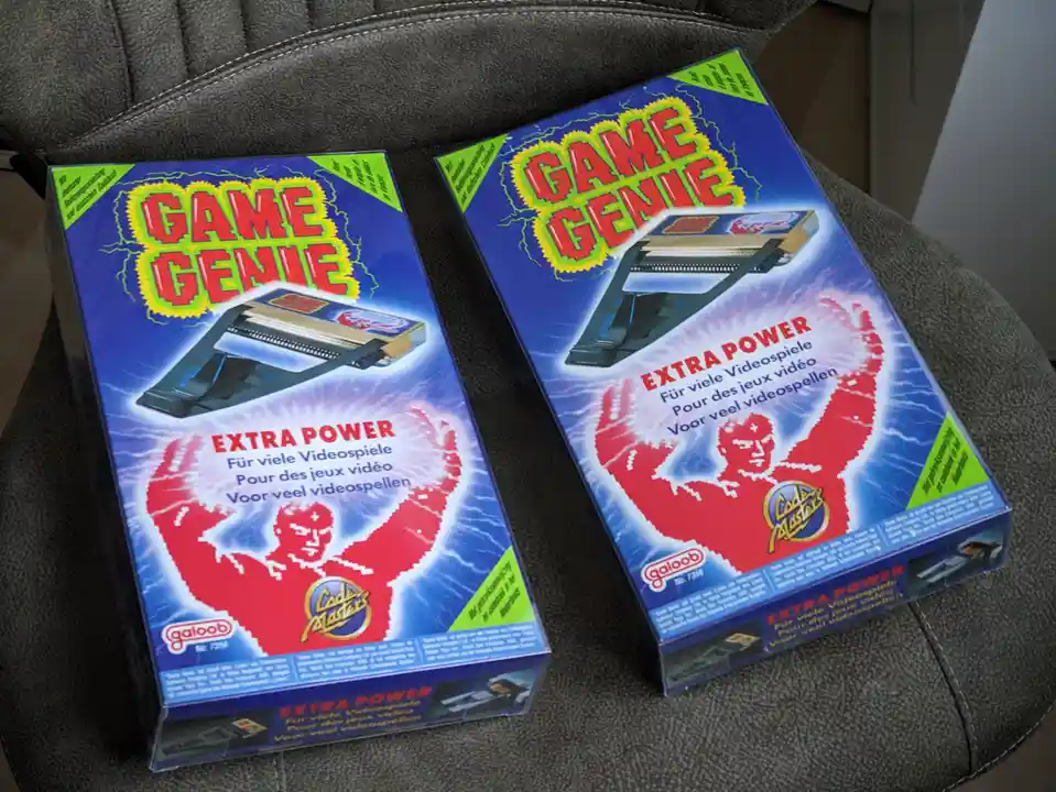 Two identical Game Genie game cheat device boxes sit side by side on a gray fabric chair. Both boxes feature the same…