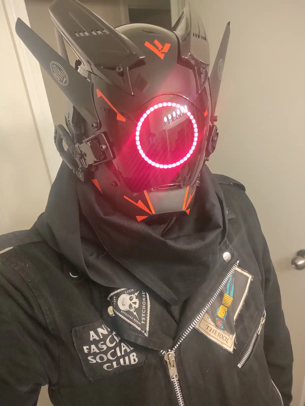 A person wearing a futuristic black helmet with glowing red LED eyes and orange accents, paired with a black jacket adorned…