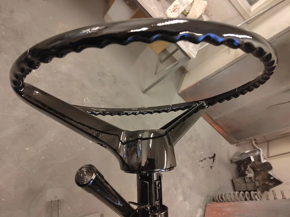 A close-up photo shows a freshly painted black steering wheel with a glossy finish, featuring a wavy, scalloped edge design…