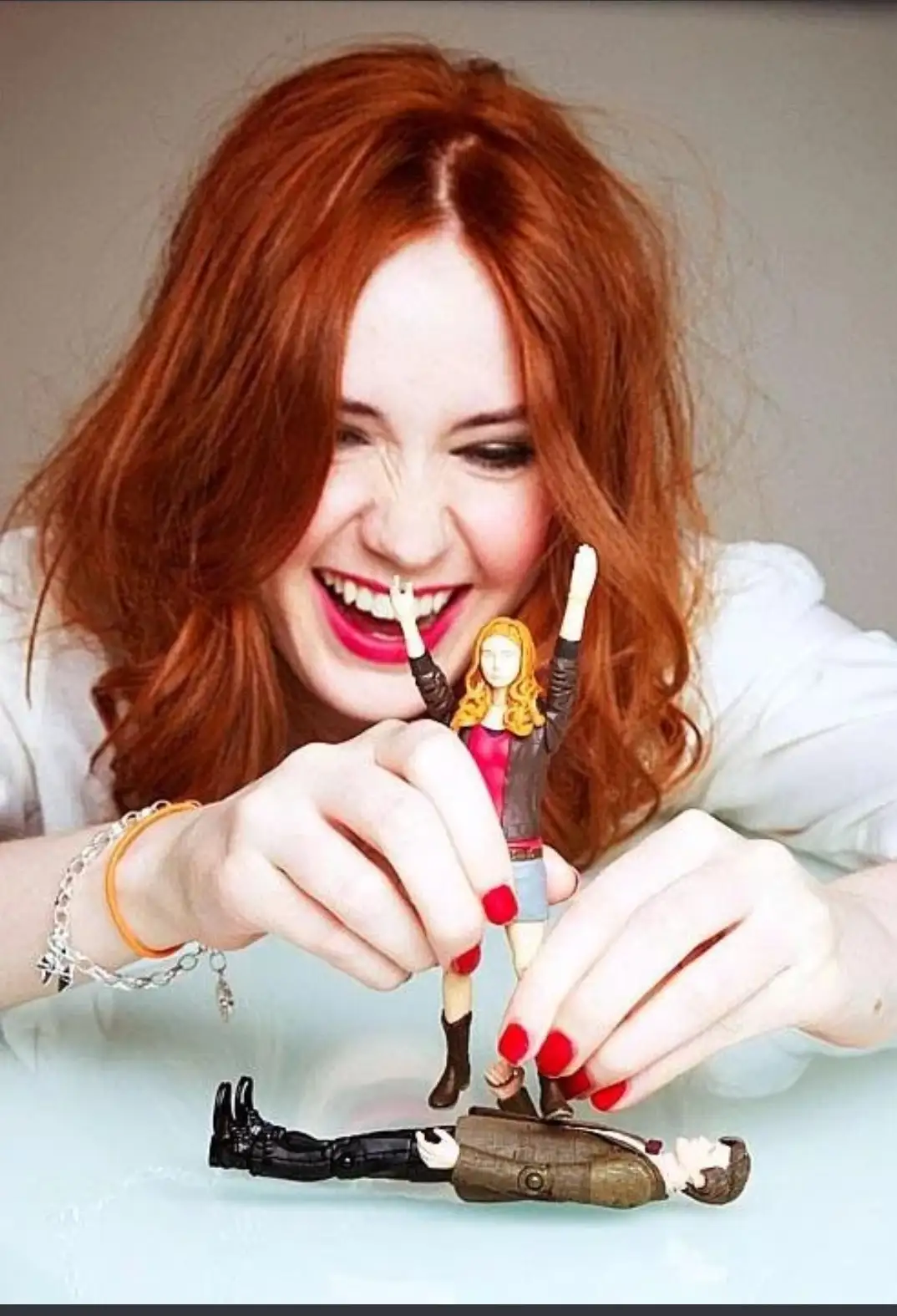 A woman with red hair and bright red fingernails holds a small figurine of a girl with blonde hair, arms raised in triumph…